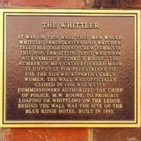 A mounted plaque titled 'The Whittler,' describing the historical significance of a local gathering spot known as Buzzard’s Roost. The plaque details how men used to whittle, trade knives and watches, tell tall tales, and chew tobacco on this wall, leading to sidewalk congestion before its official closure in 1956