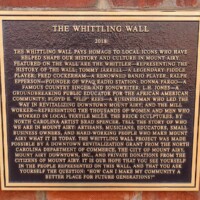 A commemorative plaque titled 'The Whittling Wall,' mounted on a brick wall and dated 2018. The text honors key figures who shaped Mount Airy’s history and culture, including musicians, educators, business leaders, and mill workers. It details the brick sculptures by North Carolina artist Brad Spencer, as well as a separate Tommy Jarrell sculpture by Mount Airy sculptor William P. Rowe, inviting visitors to reflect on the town’s rich legacy.