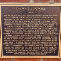 A commemorative plaque titled 'The Whittling Wall,' mounted on a brick wall and dated 2018. The text honors key figures who shaped Mount Airy’s history and culture, including musicians, educators, business leaders, and mill workers. It details the brick sculptures by North Carolina artist Brad Spencer, as well as a separate Tommy Jarrell sculpture by Mount Airy sculptor William P. Rowe, inviting visitors to reflect on the town’s rich legacy.