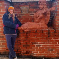 A life-sized brick sculpture of a seated musician, seamlessly integrated into the brick wall. The figure holds a guitar, leaning slightly forward as if in mid-performance. A plaque mounted above the sculpture provides historical context, though the text is not fully legible in the image. A person wearing a colorful hat, glasses, a dark jacket, purple gloves, and jeans stands beside the sculpture, adding a personal touch and sense of scale to the scene.