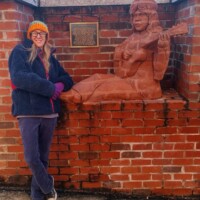 A life-sized brick sculpture of a seated musician, seamlessly integrated into the brick wall. The figure holds a guitar, leaning slightly forward as if in mid-performance. A plaque mounted above the sculpture provides historical context, though the text is not fully legible in the image. A person wearing a colorful hat, glasses, a dark jacket, purple gloves, and jeans stands beside the sculpture, adding a personal touch and sense of scale to the scene.