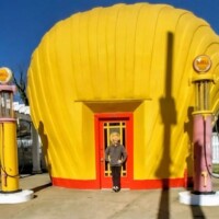 A whimsical roadside landmark: a bright yellow seashell-shaped Shell gas station with a bold red entrance, framed by retro gas pumps in yellow and pink
