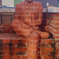A striking brick sculpture of a seated man, seamlessly integrated into a brick wall. The figure, dressed in a suit with one leg crossed over the other, conveys a relaxed yet contemplative pose. A plaque beside the sculpture identifies it as the work of Brad Spencer, created in 2015.