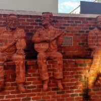 Three life-sized brick sculptures of musicians seamlessly integrated into a brick wall, each holding a different instrument—two with guitars and one with a banjo. The textured brick material makes the figures appear as if emerging from the wall itself, creating a striking, immersive effect. Two plaques mounted between the sculptures provide historical or artistic context for the installation.