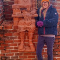 A life-sized brick sculpture of a seated man embedded into the brick wall, holding a book in one hand and a basket in the other. The reddish-brown material blends seamlessly with the surrounding architecture, creating a sculptural relief effect. A person wearing a dark jacket and purple gloves stands nearby, adding a sense of scale to the installation. In the background, a vehicle is visible above the wall, providing context to the outdoor setting.
