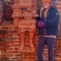 A life-sized brick sculpture of a seated man embedded into the brick wall, holding a book in one hand and a basket in the other. The reddish-brown material blends seamlessly with the surrounding architecture, creating a sculptural relief effect. A person wearing a dark jacket and purple gloves stands nearby, adding a sense of scale to the installation. In the background, a vehicle is visible above the wall, providing context to the outdoor setting.