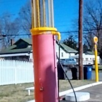 A retro Shell gas pump in soft pink with bold yellow highlights, featuring a cylindrical design and an attached hose, set against a white fence and a house under a clear blue sky.