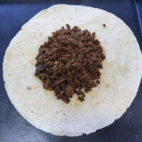 Large flour tortilla laid flat with a mound of seasoned ground beef in the center, ready for layering and folding.