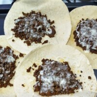 Four flour tortillas layered with seasoned beef and warm queso, arranged on a baking tray mid-assembly.