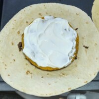 Flour tortilla with a circular tostada shell topped with sour cream placed in the center, ready for layering in a homemade Crunchwrap.