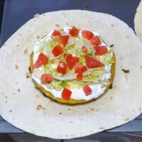 Tortilla topped with shredded lettuce, diced tomatoes, and a dollop of sour cream, partway through assembling a homemade Crunchwrap