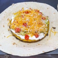 Tortilla topped with shredded lettuce, diced tomatoes, and a dollop of sour cream, partway through assembling a homemade Crunchwrap.