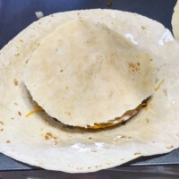 Tortilla folded over shredded cheese and seasoned beef, partially sealed in preparation for pan frying a homemade Crunchwrap.