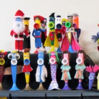 Row of colorful handmade kazoo figures displayed on a shelf—featuring characters like Santa, a police officer, a scientist, and other playful creations made from paper, fabric, and plastic. Each figure is uniquely decorated with googly eyes, pipe cleaners, and vibrant accessories. Quirky, charming, and deeply kazoo-coded