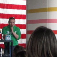 A speaker in a green shirt stands at a microphone in front of a giant American flag backdrop made from red, white, and blue plastic cups. A multi-colored striped column stands nearby, and children are visible in the audience foreground. Loud, proud, and fully kazoo-core
