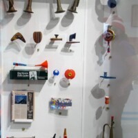 Display case filled with small rocket parts, fins, nose cones, tools, space-themed models, a miniature satellite, and a trumpet-like object—arranged on a white background with blue stripes. A kazoo-adjacent tribute to rocketry and sonic experimentation.
