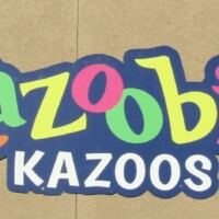 Kazooie Kazoos sign in bright, playful lettering—‘Kazooie’ spelled in multicolor cartoon-style letters above bold white ‘Kazoos,’ mounted on a beige wall with overhead spotlights. Where whimsy goes full volume.