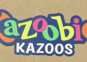 Kazooie Kazoos sign in bright, playful lettering—‘Kazooie’ spelled in multicolor cartoon-style letters above bold white ‘Kazoos,’ mounted on a beige wall with overhead spotlights. Where whimsy goes full volume.