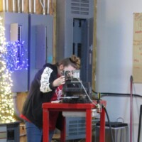 Person in a black and white jacket operates machinery atop a red table, surrounded by electrical panels, wall writing, and workshop gear. Kazoo manufacturing meets mystery lab chic.