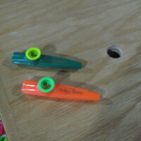 Two colorful plastic kazoos—one green with a yellow resonator, the other orange with a green resonator—resting on a wooden surface with a nearby hole and a scattering of other kazoo colors in the bottom corner. Childhood chaos, staged beautifully.