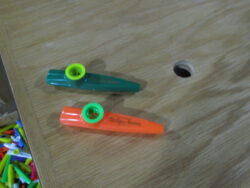 Two colorful plastic kazoos—one green with a yellow resonator, the other orange with a green resonator—resting on a wooden surface with a nearby hole and a scattering of other kazoo colors in the bottom corner. Childhood chaos, staged beautifully.