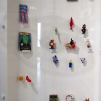 Glass display case filled with miniature kazoo-themed figures, trinkets, and oddities—each arranged against a white backing. The collection includes clown heads, toy animals, kazoo hybrids, and various whimsical characters. A pocket-sized tribute to musical mayhem.