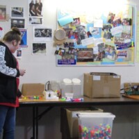Person in a black and white jacket stands at a table cluttered with craft items, boxes, and colorful supplies. Behind them, a bulletin board is packed with handwritten notes, photos, and flyers, including a bright yellow sheet with large handwriting. A shrine to organization, panic, and possible kazoo conspiracies.