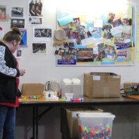 Person in a black and white jacket stands at a table cluttered with craft items, boxes, and colorful supplies. Behind them, a bulletin board is packed with handwritten notes, photos, and flyers, including a bright yellow sheet with large handwriting. A shrine to organization, panic, and possible kazoo conspiracies.