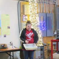 Tour guide in a red ‘Kazooie Kazoos’ shirt holding a tray, standing in a workshop decorated with string lights, posters, and kazoo-making equipment. The face of calm in a sea of kazoo chaos