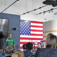 Audience seated in a room facing a screen that reads ‘Starring Kanokie Kazan’—a person stands in front of it, mid-presentation. American flag on one wall, rainbow-striped panel on the other. Lights, camera, kazoo.