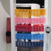 Interactive display with colorful horizontal woven strips in a vertical frame—featuring yellow, orange, pink, red, and blue bands. A pair of black headphones hangs on the right, and a wooden panel with a dark circular sensor or speaker is on the left. Whimsy meets mystery in kazoo-tech form.