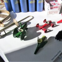 Assorted toy instruments and vehicles—including a red car, green airplanes, black toy car, miniature trumpet, and trombone—displayed with blue plastic tubes, boxed items, and scattered trinkets. The ultimate kazoo-core gift shop table.