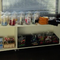 Merchandise shelf with clear jars of colorful trinkets, stacks of folded t-shirts, and lower bins full of boxed snacks, kazoo souvenirs, and impulse treasures. A rainbow mess of whimsy, sealed in plastic and hope.