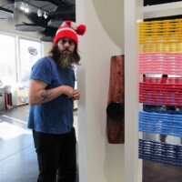Person in a red-and-white hat and boots stands next to a white interactive display filled with colorful woven bands and a wooden kazoo sculpture. Part performance art, part instruction manual. Definitely not OSHA approved