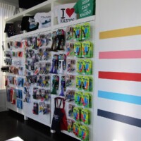 Merch wall filled with colorful kazoo-themed items including socks, accessories, tote bags, and novelty signs. Prominent signs read ‘KEEP CALM AND KAZOO’ and ‘I ♥ KAZOONE.’ Striped wall of yellow, pink, red, blue, and teal adds bold color. The fashion frontier of kazoo fandom.