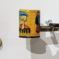 Wall-mounted kazoo sculpture made from a metal pipe and a cartoon-covered can—featuring Popeye and Wimpy illustrations—with a yellow and black ‘10’ mounted beside it. Vintage, scrappy, and weirdly charming.