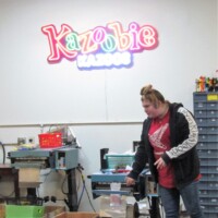 Kazooie Kazoos neon sign glowing above a busy workshop where a person in a red shirt and patterned black jacket stands at a machine. Surrounding shelves hold bins and drawers filled with supplies. A factory floor where whimsy goes professional