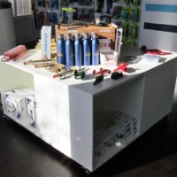 Display table at the Kazooie Kazoos gift shop featuring blue water bottles, a red toy car, green and red toy airplanes, white boxes with blue labels, and a mix of tools, trinkets, and playful chaos. Souvenirs that shout instead of whisper.