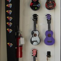 Six colorful ukuleles mounted on a white wall—ranging from black and floral to bright red and soft purple—next to a vertical strip of diamond-shaped stickers and a red fire extinguisher. Unexpectedly artsy, unapologetically kazoo-adjacent.