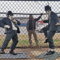 Statues of Jake and Elwood Blues from The Blues Brothers, dressed in black suits, sunglasses, and black hats, standing in front of a chain-link fence. Jake is posed adjusting his tie, while Elwood strikes a mid-dance stance. Behind them, Zach stands playfully in the background, partially visible through the fence, adding an extra layer of personality to the scene. Captured outdoors in Stedman, NC.