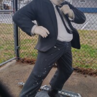 Statue of Jake Blues from The Blues Brothers, dressed in a black suit, white shirt, black tie, and black hat, posed dynamically with one hand adjusting his tie. The statue stands on a metal grate, with a chain-link fence behind it. Captured outdoors in Stedman, NC.