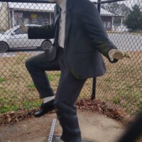 Statue of Elwood Blues from The Blues Brothers, dressed in a black suit, white shirt, black tie, black hat, and sunglasses. He is posed mid-dance, with one leg raised and arms positioned dynamically. The statue stands on a metal grate, enclosed by a chain-link fence, with a car and building in the background. Captured outdoors in Stedman, NC.