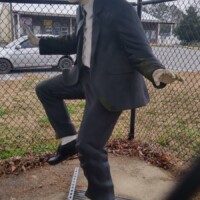 Statue of Elwood Blues from The Blues Brothers, dressed in a black suit, white shirt, black tie, black hat, and sunglasses. He is posed mid-dance, with one leg raised and arms positioned dynamically. The statue stands on a metal grate, enclosed by a chain-link fence, with a car and building in the background. Captured outdoors in Stedman, NC.
