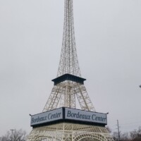 Miniature white Eiffel Tower replica standing in a commercial parking lot at Bordeaux Center in Fayetteville, North Carolina, framed by a blue sky and shopping plaza backdrop.