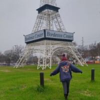 Person walking toward a white Eiffel Tower replica at Bordeaux Center in Fayetteville, North Carolina, with 'Bordeaux Center' signage visible and trees and a medical building in the background.