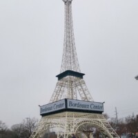 White Eiffel Tower replica at Bordeaux Center in Fayetteville, North Carolina, with a sign reading 'Bordeaux Center' across the middle and a person standing near the base, under an overcast sky.