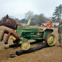 Creative roadside sculpture blending the front half of a painted horse with the back half of a green tractor, complete with yellow wheels and a mannequin posed as the driver. The sculpture sits on a wooden platform in a grassy clearing, with a person standing beside it and trees under a cloudy sky in the background