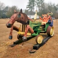 Sculpture of a hybrid creature with the front half resembling a painted horse and the rear half built from a green tractor, with yellow wheels and a seated mannequin posed as the driver. A person stands beside it for scale, with trees and buildings in the background.