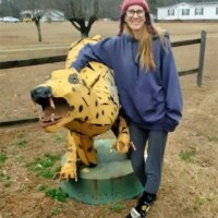 Large yellow leopard sculpture with black spots and an open mouth, displayed in a grassy area near a fenced boundary. A person stands beside it, resting one arm on the leopard, with trees and a white building in the background.