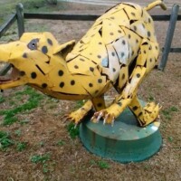 Metal sculpture of a leaping yellow cheetah with black spots, crafted from welded parts and mounted on a green circular base in an outdoor setting with grass and a wooden fence.